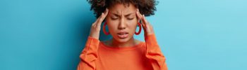 Photo of frustrated young Afro American woman has headache keeps hands on temples suffers unbearable migraine after noisy party wears orange sweater poses indoor isolated on blue background.